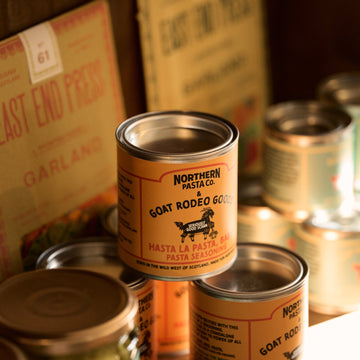 Cans of Northern Pasta Co. goat rodeo gold pasta on a wooden surface with a blurred background.