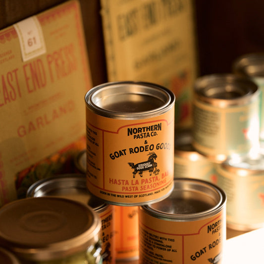 Cans of Northern Pasta Co. goat rodeo gold pasta on a wooden surface with a blurred background.