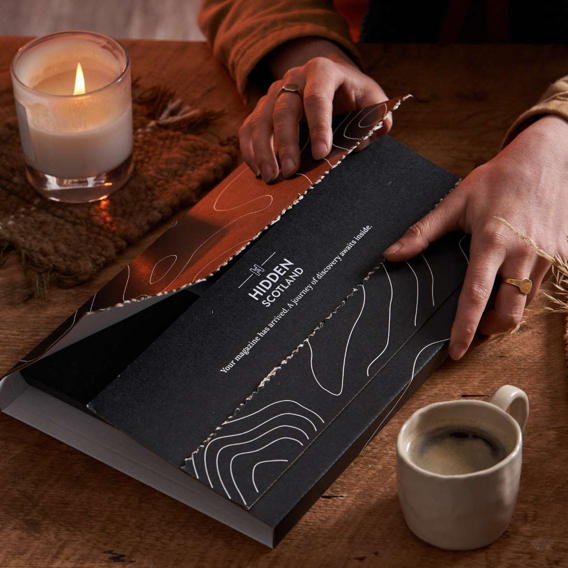 Person opening a black box with 'Hiddin' Scotland' branding on a wooden table with a lit candle and cup of coffee.